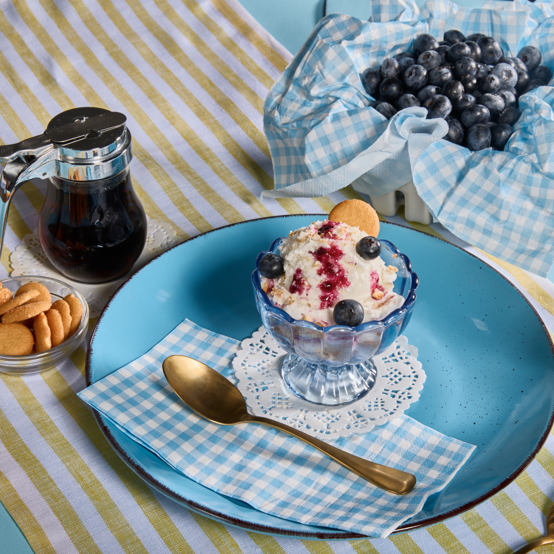 Blueberry coconut milk ice cream swirled with berry sauce, topped with fresh blueberries and wafer cookies, served in glass cups with syrup, cookies, and a basket of blueberries nearby.