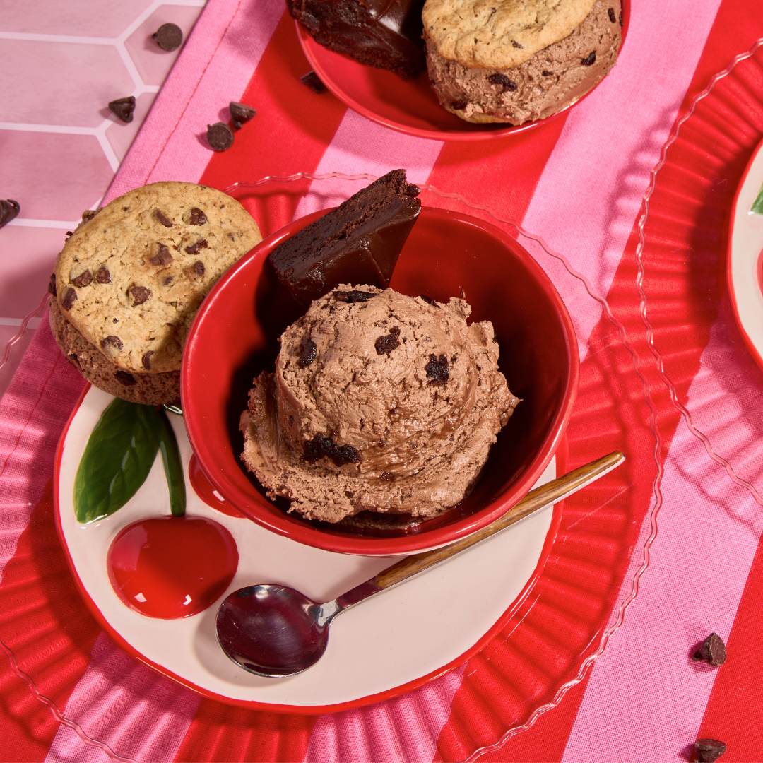 Double chocolate brownie ice cream topped with chocolate chunks, served with brownies and chocolate chip cookies on a pink dessert table.