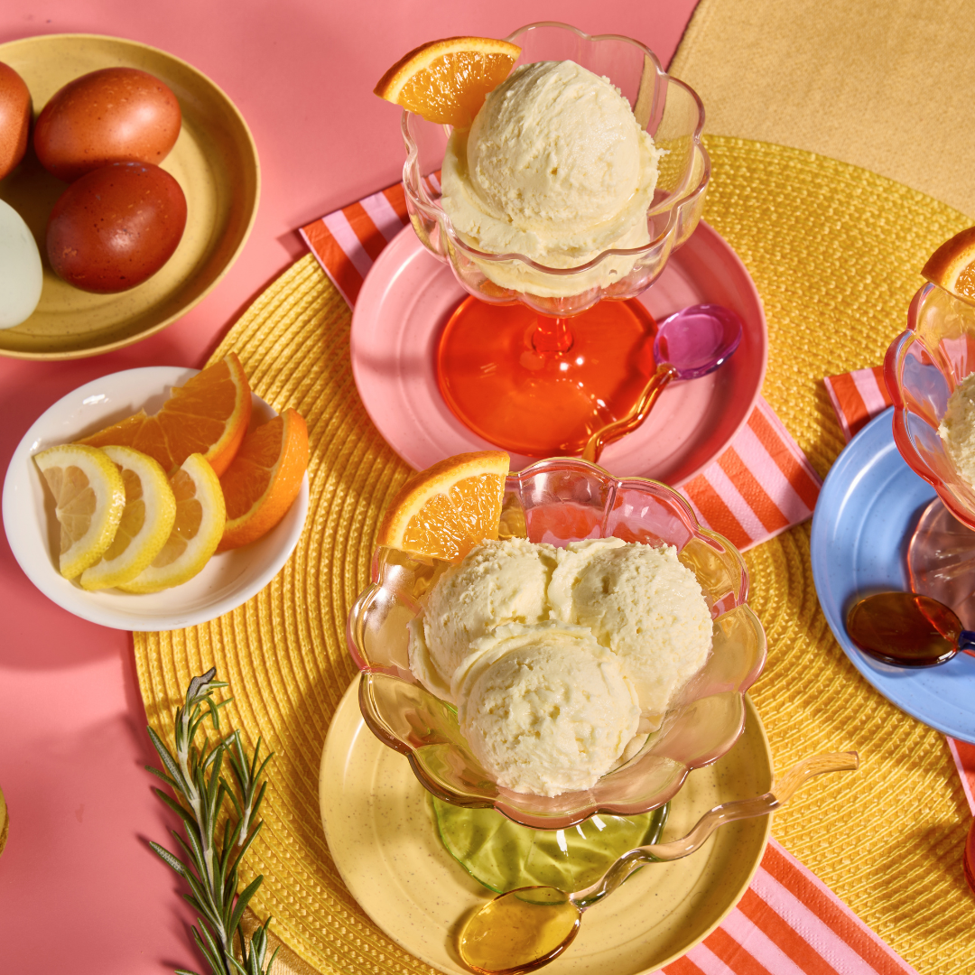 Three scoops of classic custard served in colorful glass dessert bowls with citrus slices and spoons on a bright pastel table.