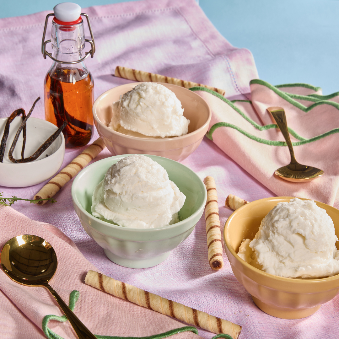 Three pastel bowls filled with scoops of classic vanilla ice cream sit on a soft pink cloth, surrounded by vanilla beans, wafer rolls, a small glass bottle of vanilla extract, and gold spoons, styled with pink flowers on a light blue background.
