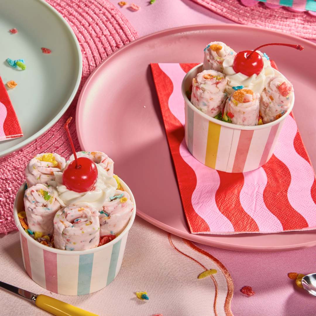 Fruity Pebbles rolled ice cream topped with whipped cream, cherries, and colorful cereal pieces in striped dessert cups.