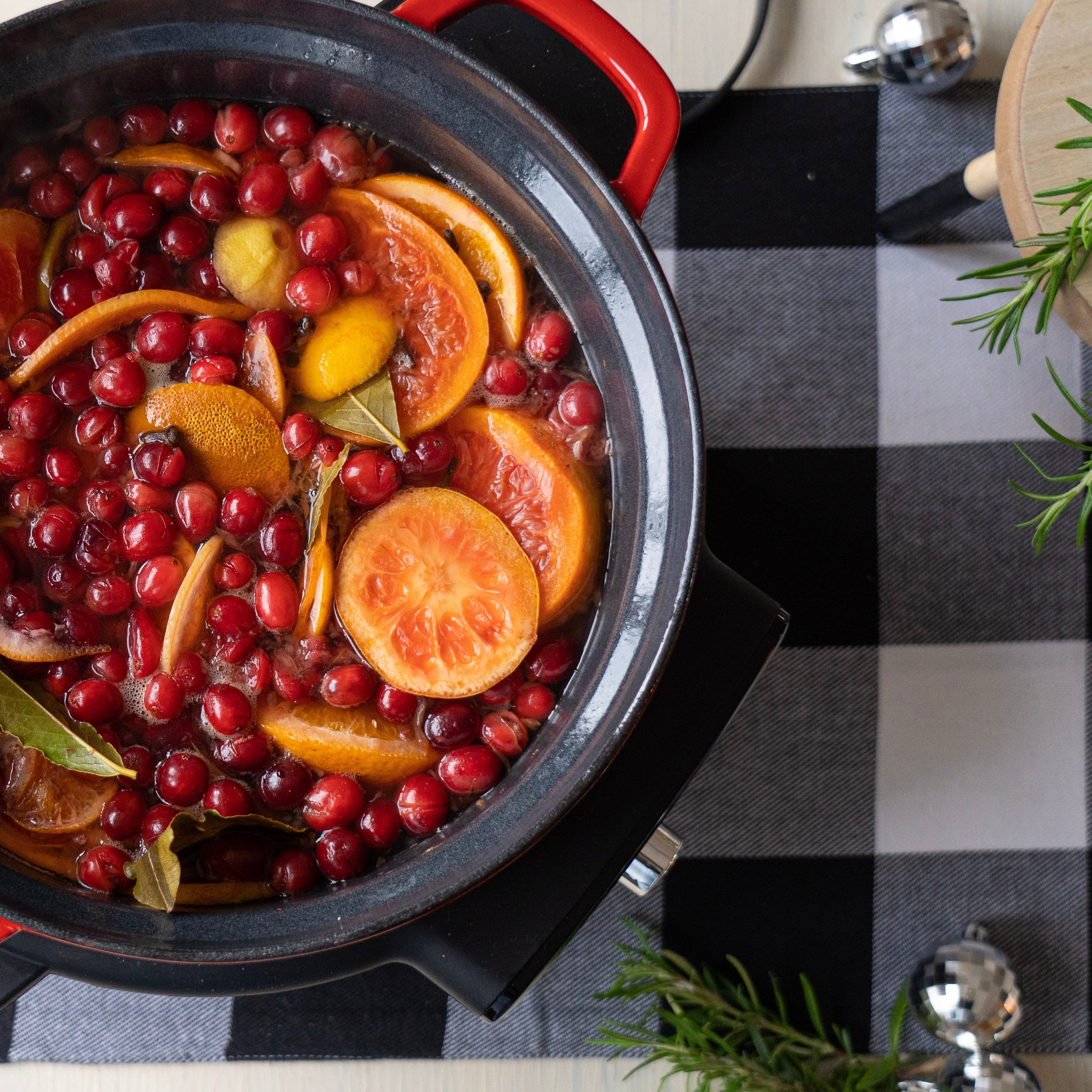 Overhead view of a simmering holiday potpourri in a black pot with red handles, filled with bright red cranberries, sliced oranges, lemon rounds, and bay leaves floating in warm liquid.