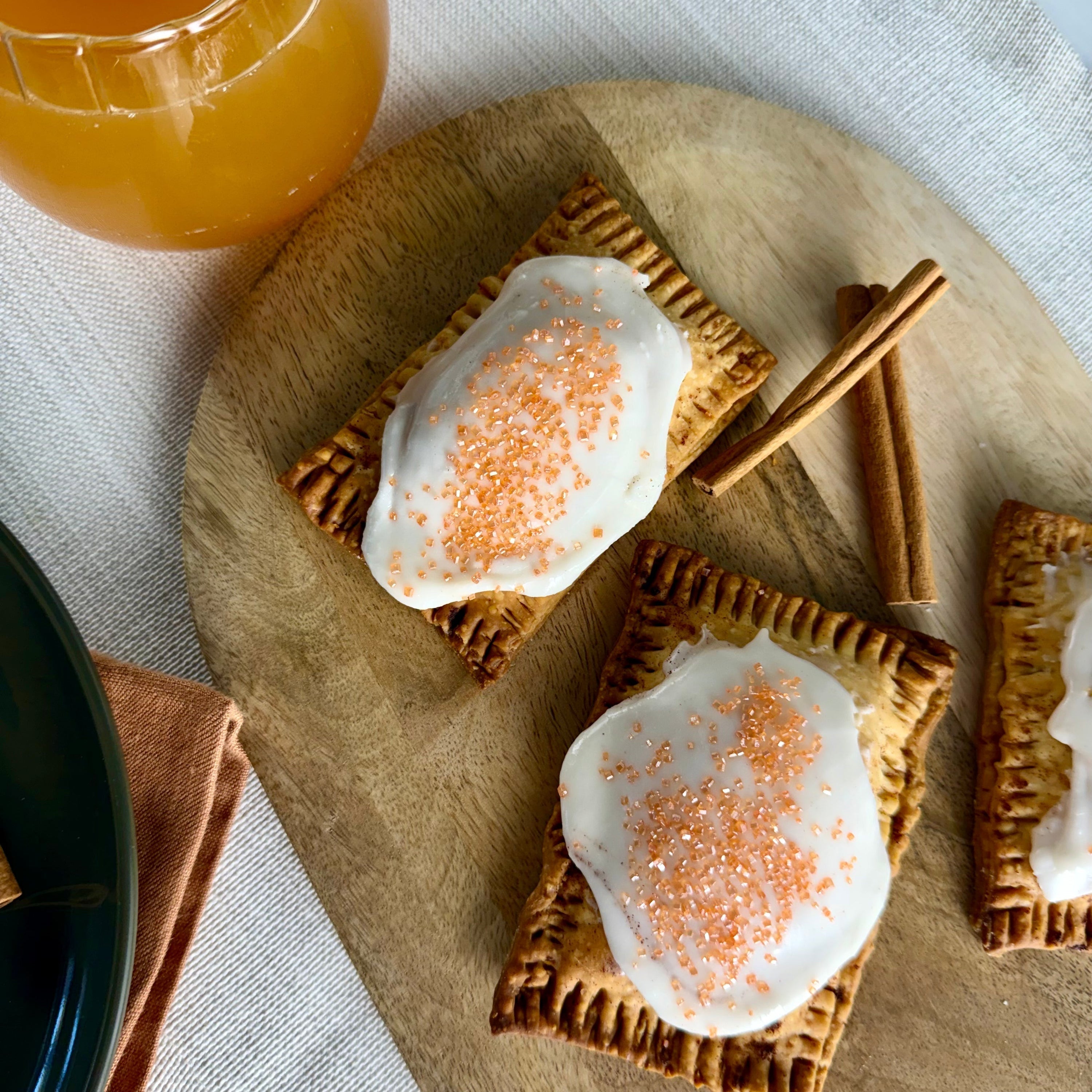 Homemade pumpkin toaster pastries with white icing and orange sprinkles on a wooden board, with cinnamon sticks and a glass of apple cider beside them.