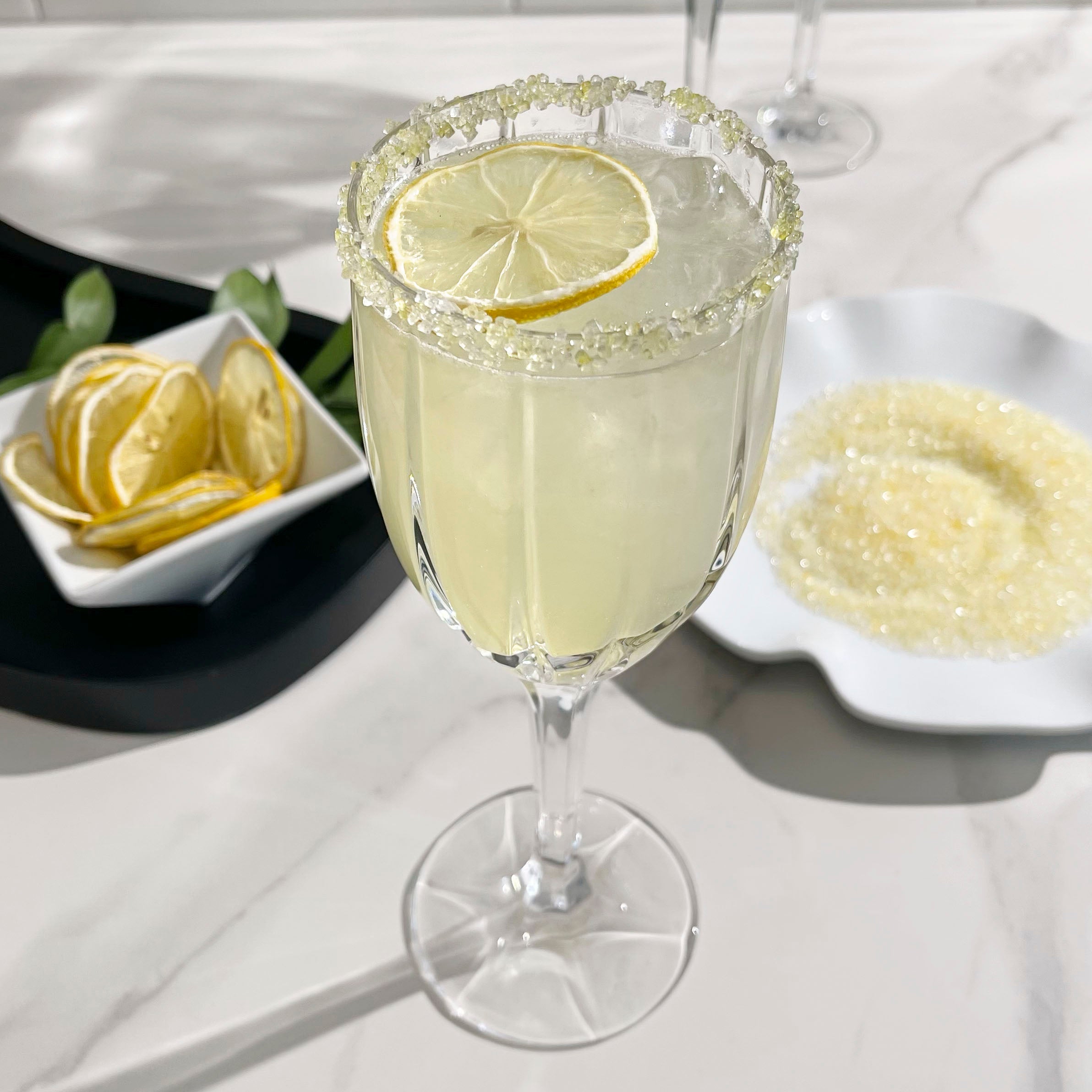 Limoncello and sage spritz in a clear stemmed glass with a sugared rim and lemon slice garnish, set on a light countertop.