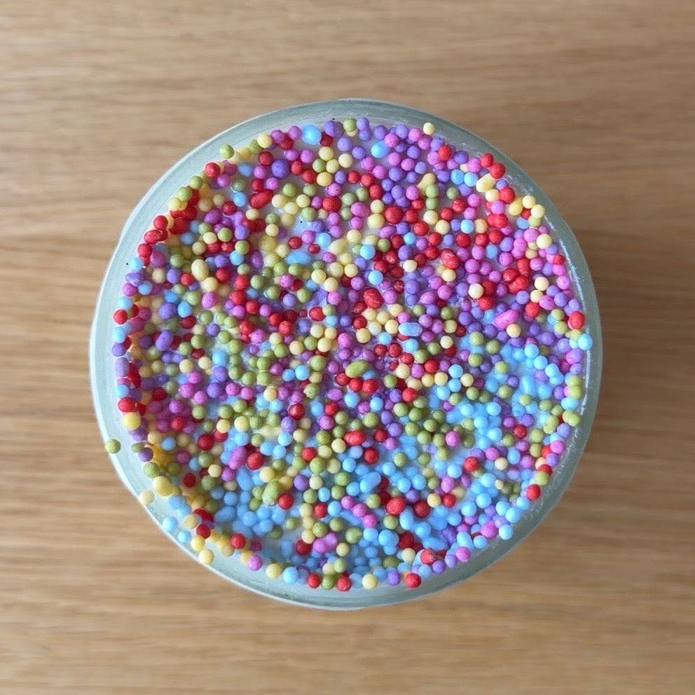 Overhead view of a mini vanilla ice cream dot cake served in a clear glass. The top layer of vanilla ice cream is coated with colorful rainbow nonpareil sprinkles in shades of pink, purple, blue, green, yellow, and red. 