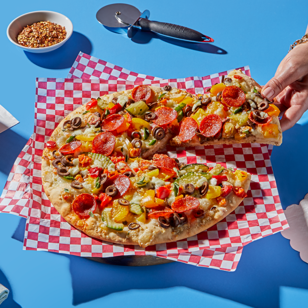 Hand lifting a slice from a vegetable and pepperoni pizza on red-and-white checkered paper over a blue tabletop, with a pizza cutter and a small bowl of crushed red pepper nearby.