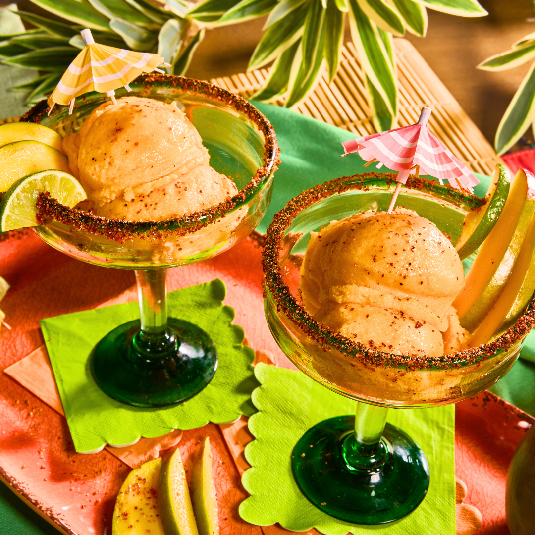 Two margarita-style glasses filled with mango lime Tajín sorbet, garnished with lime wedges and chili-lime rims.