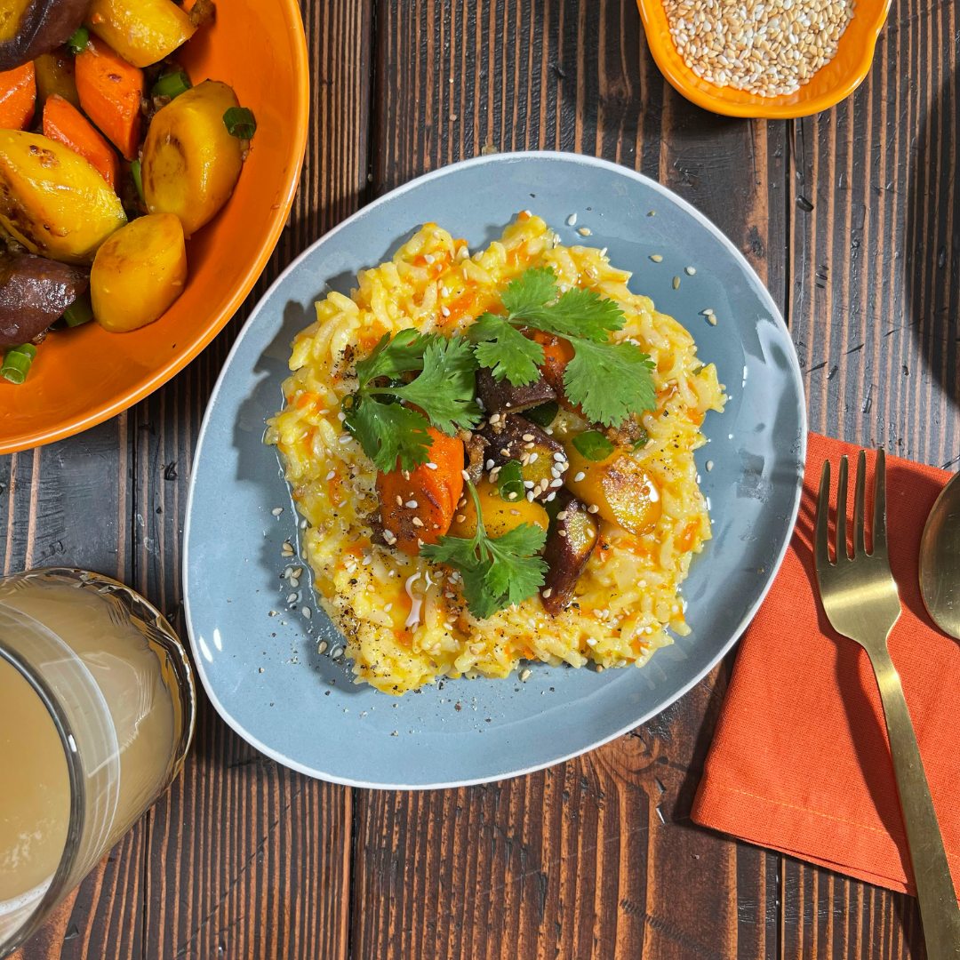 A plate of carrot risotto made with arborio rice, grated orange carrots, miso, butter, and Parmesan, topped with sautéed tri-color carrots glazed in miso butter with ginger and scallions. The dish is garnished with sesame seeds and fresh cilantro leaves.