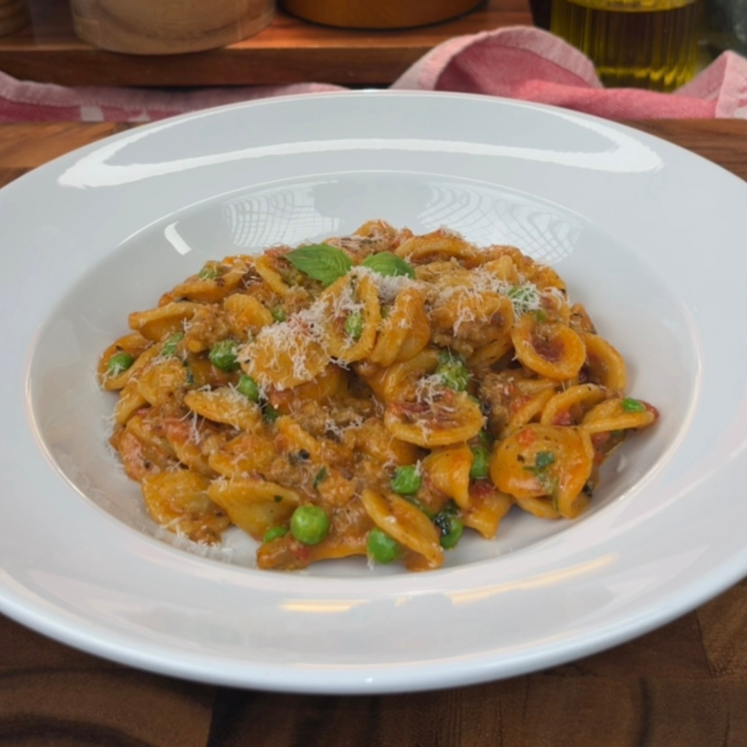 A white bowl filled with creamy orecchiette pasta mixed with Italian sausage and green peas, topped with grated Parmesan and a small basil leaf.