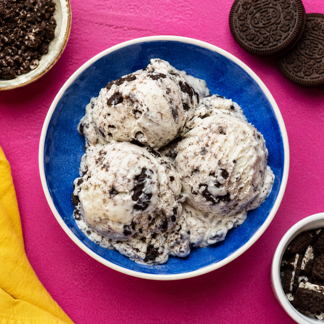 Three scoops of cookies and cream ice cream in a blue bowl on a bright pink background with Oreo cookies, crumbs, and a yellow napkin.