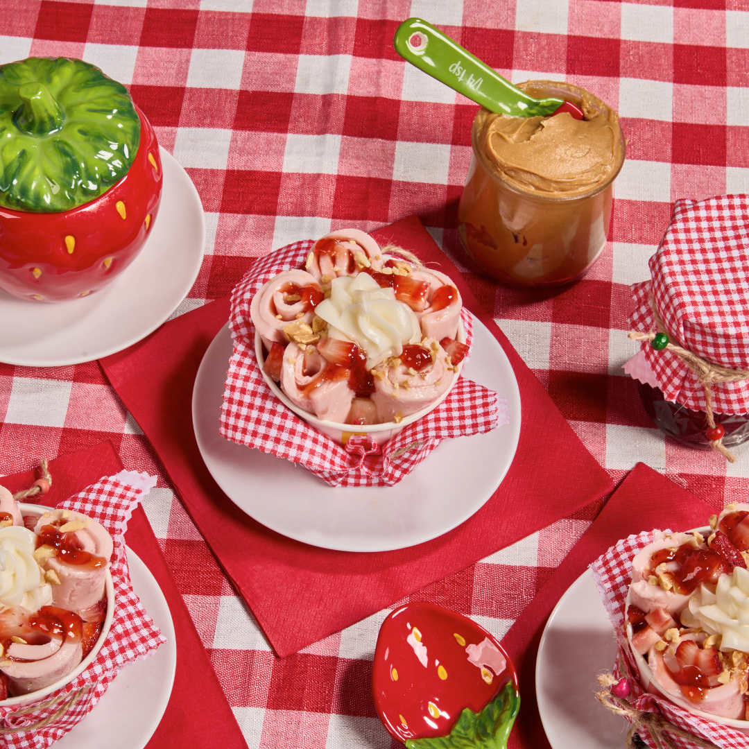 PB&J rolled ice cream topped with strawberries, jam, peanuts, and whipped cream, served on a red gingham picnic table.