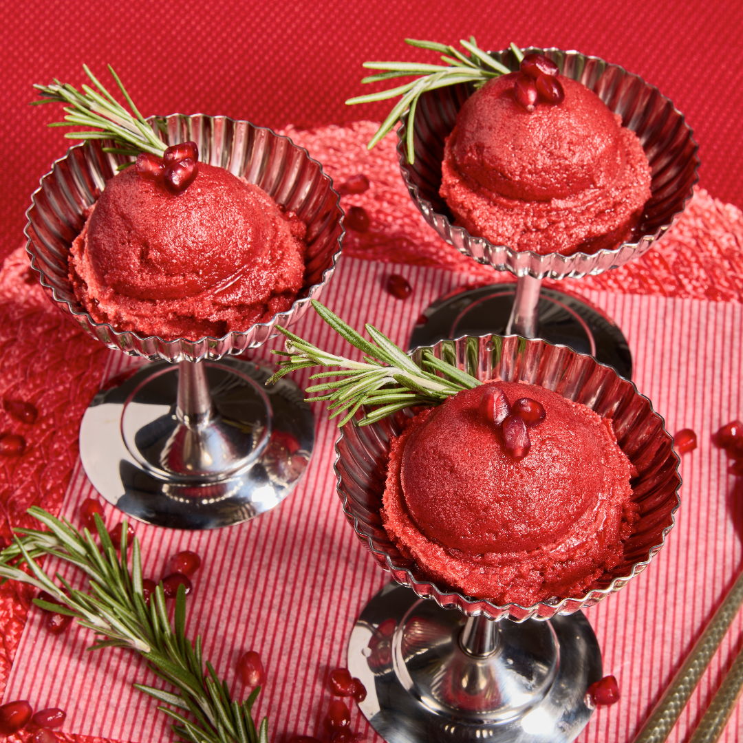 Three silver dessert cups filled with vibrant pomegranate cranberry sorbet, topped with rosemary sprigs and pomegranate seeds, styled on a rich red backdrop with scattered arils.