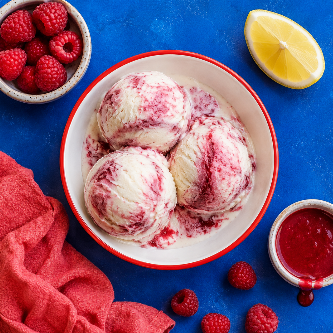 Top-down photo of three raspberry lemon gelato scoops with pink berry swirls in a white bowl on a bright blue surface, surrounded by fresh raspberries, lemon, raspberry sauce, and a coral napkin.