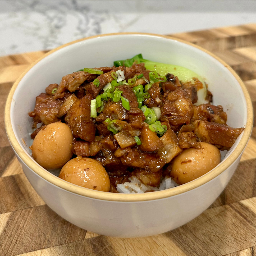 A bowl of Taiwanese Braised Pork Belly served over white rice, topped with glossy, caramelized pork pieces, halved canned quail eggs, and chopped green onions. A few pieces of bright green bok choy sit along the side of the bowl.