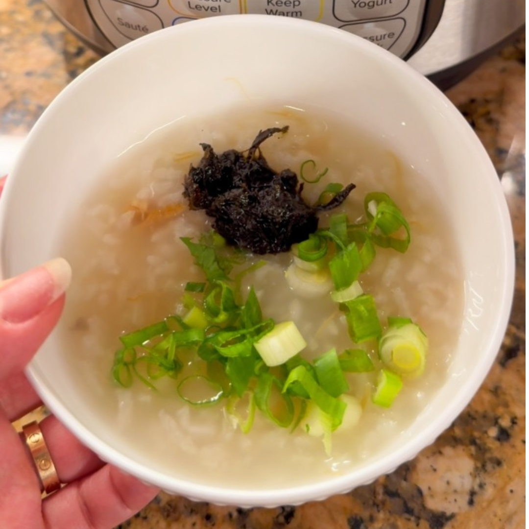 A white bowl of Chinese porridge topped with chopped green onions and a small portion of dark preserved vegetable. The bowl is held in one hand, with an Instant Pot® control panel visible in the background.