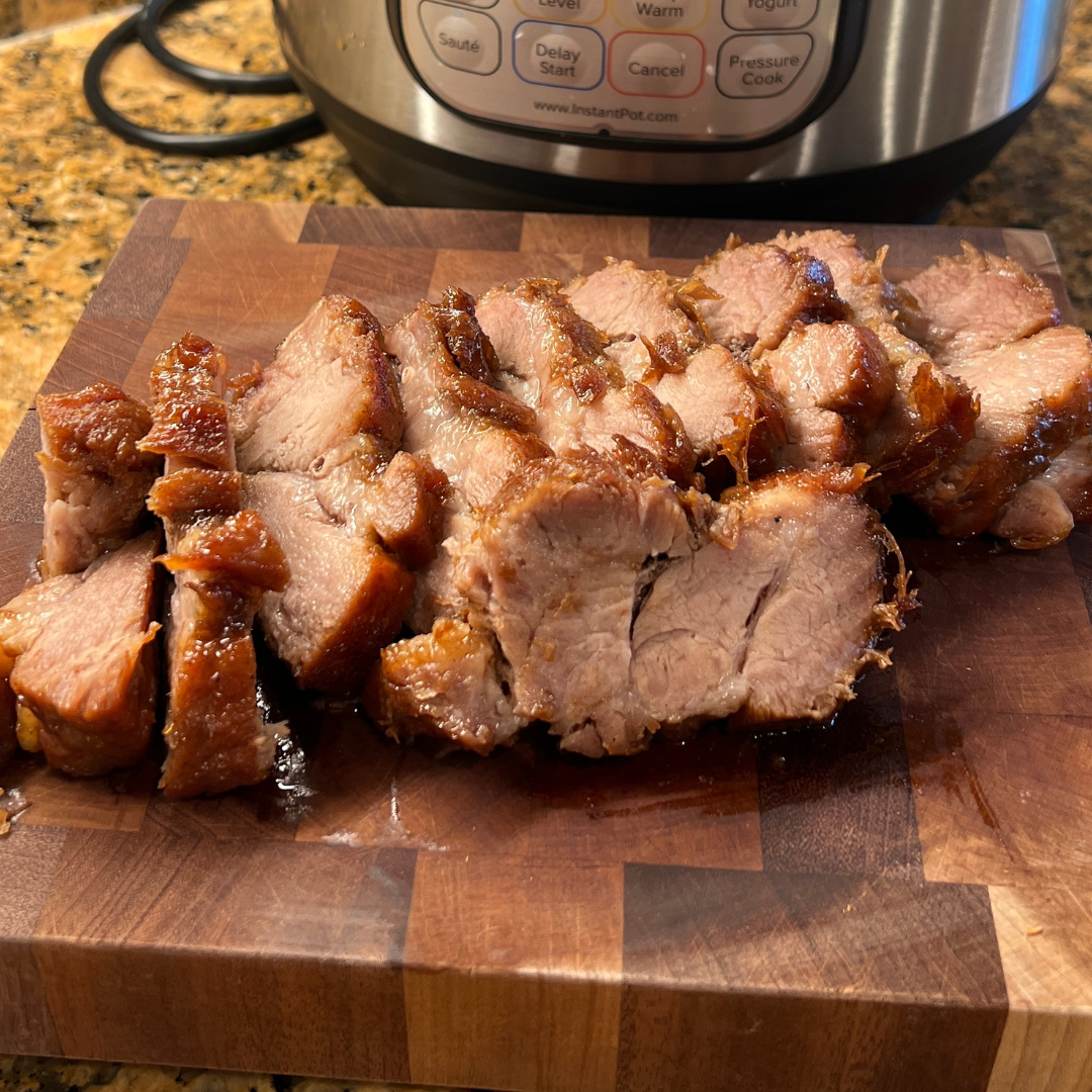 Sliced Char Siu pork arranged on a wooden cutting board. The meat is glazed with a glossy, caramelized honey sauce, showing a tender, juicy interior with slightly crisp edges. An Instant Pot® control panel is visible in the background.
