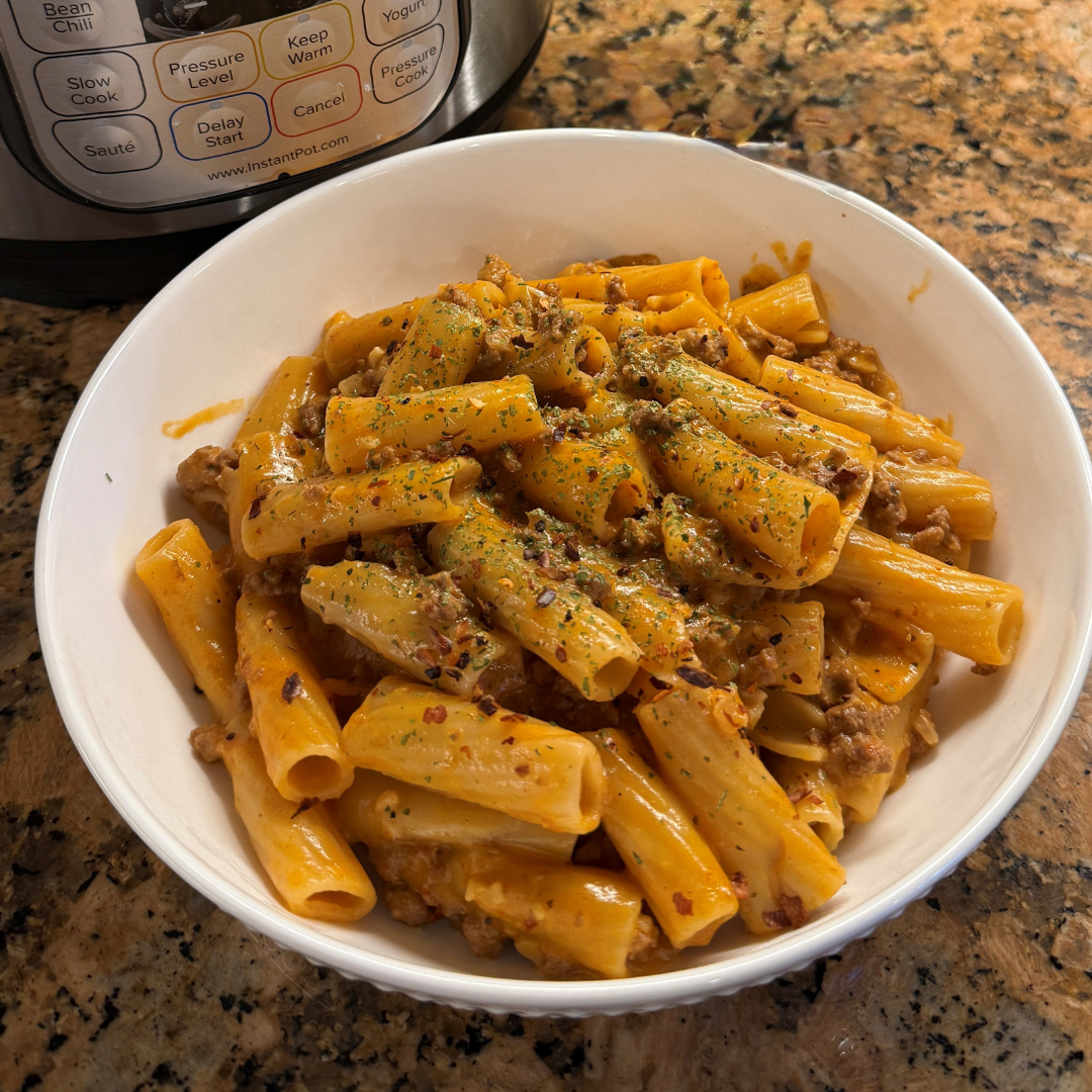 A white bowl filled with rigatoni pasta coated in creamy vodka sauce, mixed with ground meat and topped with dried herbs and red pepper flakes. The pasta is rich and golden-orange in color, with an Instant Pot® visible in the background.