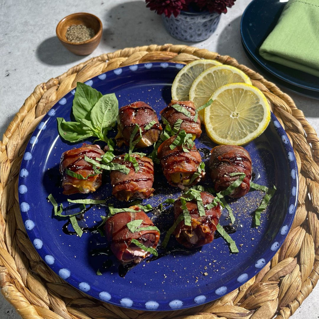 A deep blue plate holds several prosciutto-wrapped figs stuffed with goat cheese, drizzled with balsamic glaze and sprinkled with fresh basil ribbons.