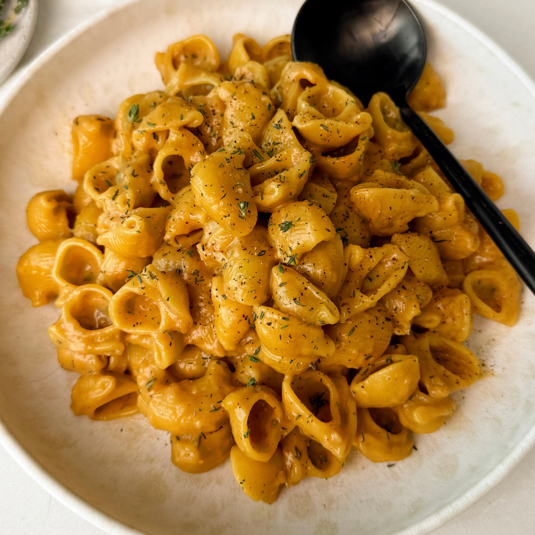 Close-up of creamy French onion pasta shells coated in a rich golden-brown sauce, garnished with specks of herbs and black pepper, served in a white bowl with a black spoon resting beside the pasta.