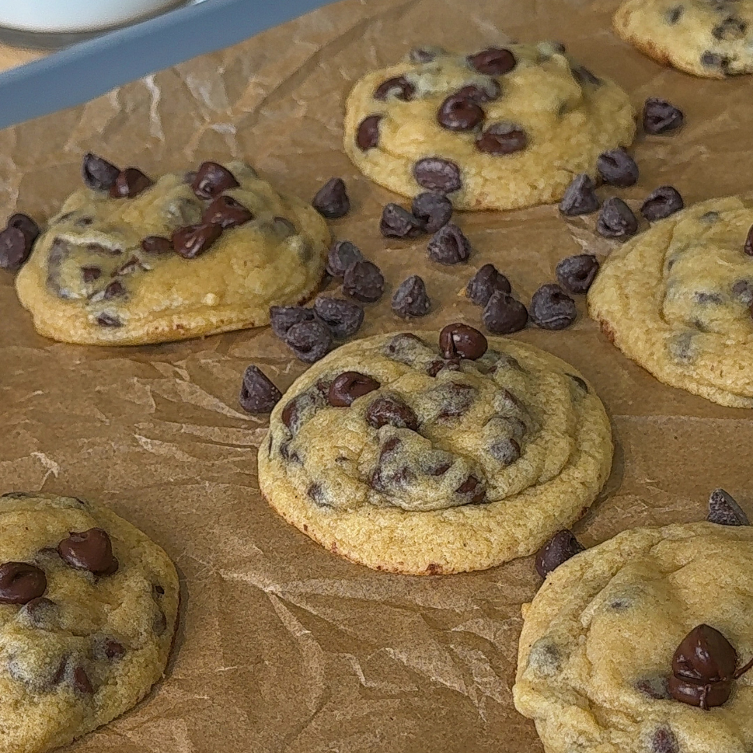 Close-up of soft and chewy chocolate chip cookies baked on crinkled parchment paper, with golden edges, pale centers, and melted chocolate chips visible throughout. Several loose chocolate chips are scattered between the cookies.