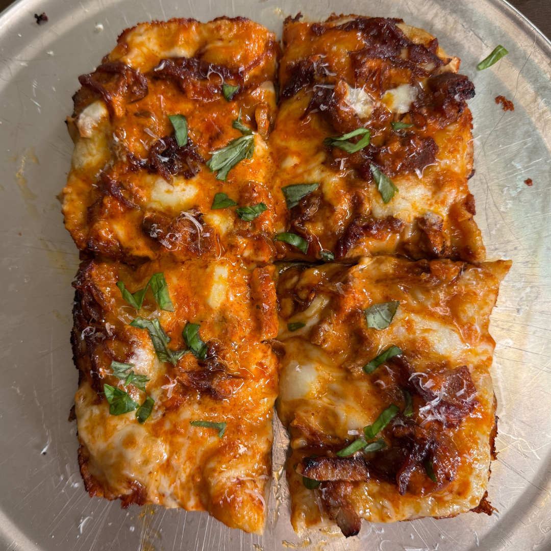 Close-up of a rectangular pizza cut into four slices, topped with melted mozzarella, rich red tomato sauce, caramelized pork belly pieces, and fresh basil scattered on top