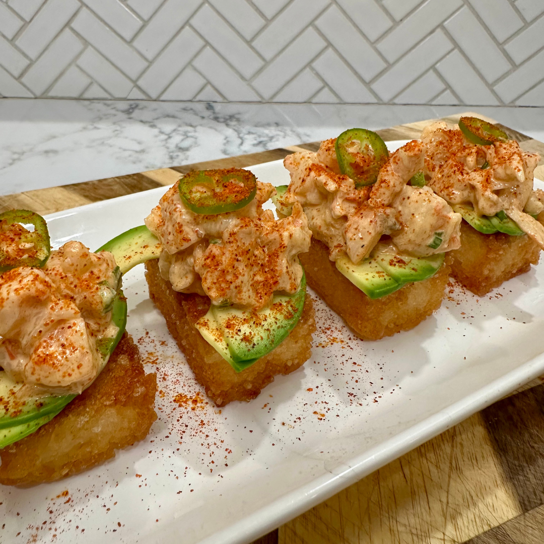 Spicy shrimp crispy rice bites topped with avocado slices, creamy shrimp mixture, sliced jalapeños, and a dusting of chili powder, served on a rectangular white plate with a tiled backsplash in the background.