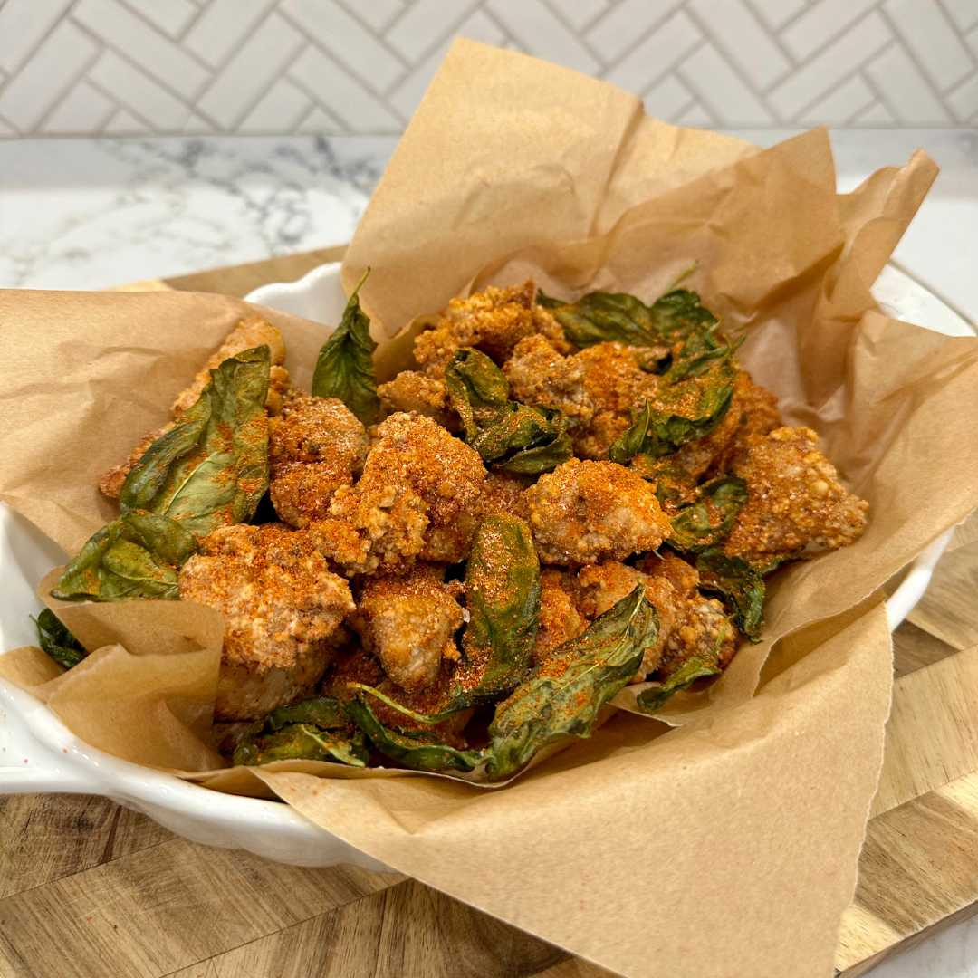 Taiwanese Popcorn Chicken served in a paper-lined bowl, topped with crispy Thai basil leaves and a dusting of chili powder, set on a wooden board with a white tiled background.
