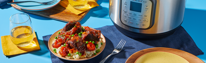 A plated meal of braised beef short ribs with carrots, peas, and mashed potatoes sits beside an Instant Pot® Duo™ Crisp multi-cooker on a blue table, with a glass of white wine, fork, and empty plate nearby.