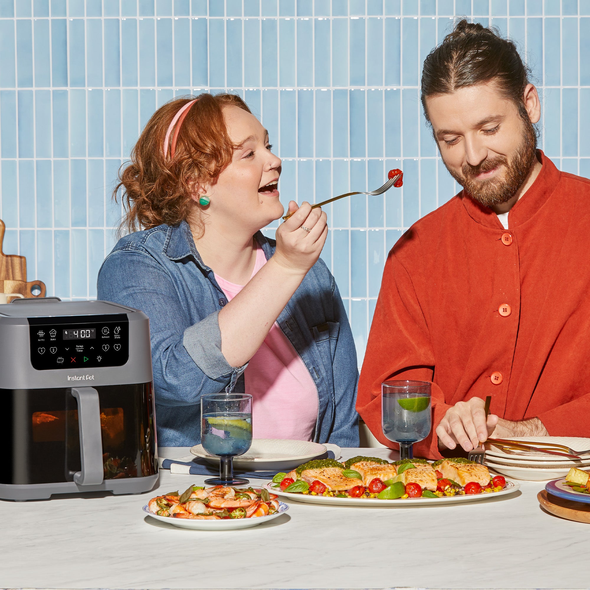 Two people seated at a table with the air fryer beside them. A woman in a denim shirt playfully feeds a forkful of food to a man in a red shirt. Plates of colorful roasted salmon, vegetables, and drinks are on the table.