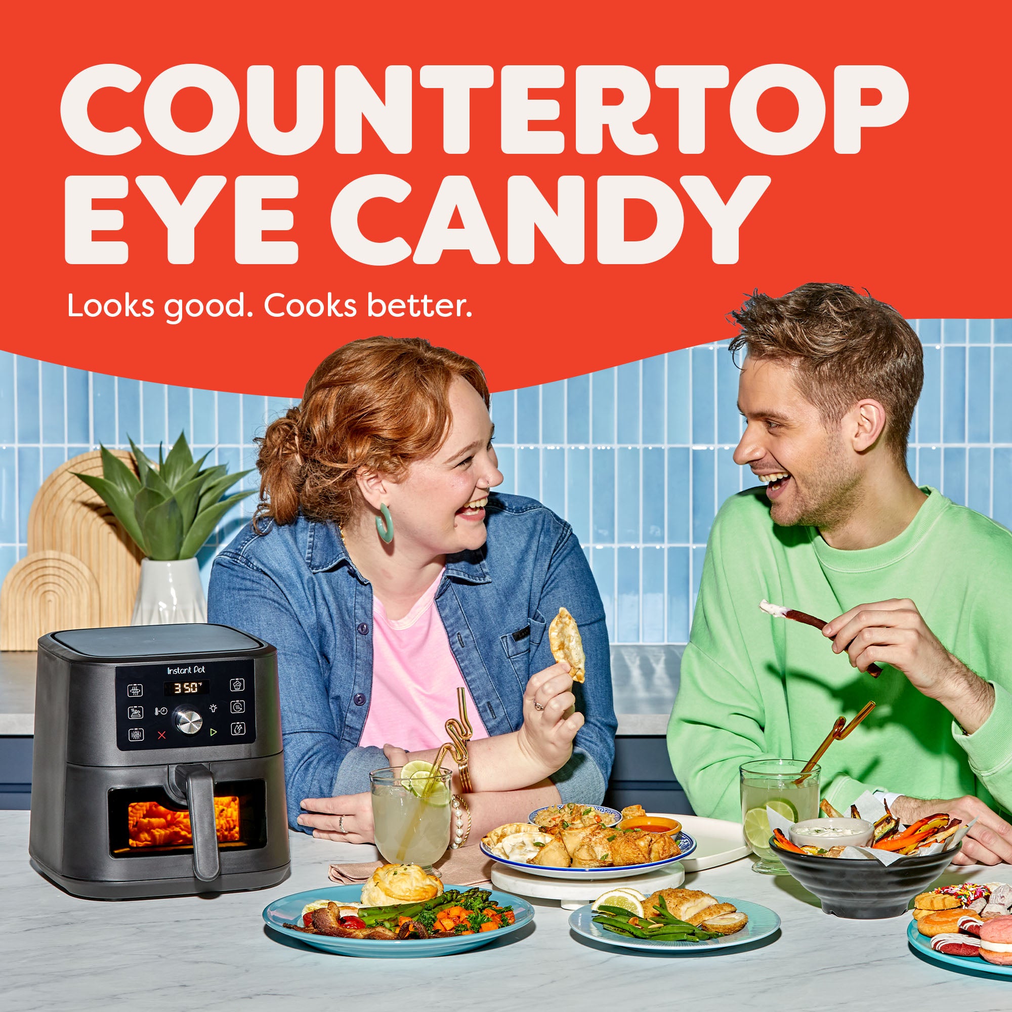 Two smiling people sit at a kitchen counter enjoying a variety of colorful meals, with the Instant Pot® 4QT Vortex® Plus Mini Air Fryer in Black on the counter beside them. The appliance shows a 350°F reading. Text above reads “Countertop Eye Candy – Looks good. Cooks better.” A potted plant and bright tiled backsplash complete the vibrant kitchen scene.
