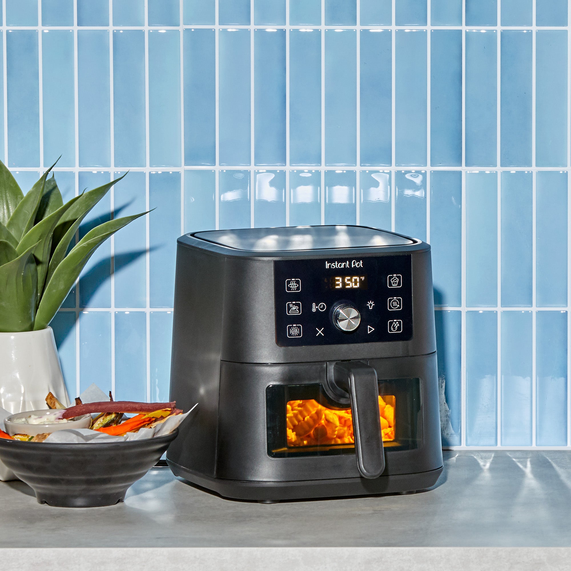 The Instant Pot® 4QT VORTEX® Plus Mini Air Fryer in Black sits on a kitchen countertop in front of a glossy blue tile backsplash, cooking crinkle-cut fries through its viewing window. A bowl of colorful veggie fries with dip is placed beside it.