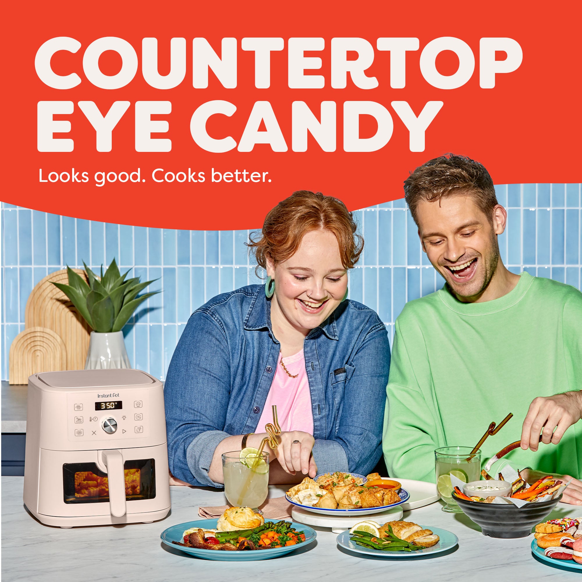 Two smiling people sit at a kitchen counter enjoying a variety of colorful meals, with the Instant Pot® 4QT Vortex® Plus Mini Air Fryer in Rosewater on the counter beside them. The appliance shows a 350°F reading. Text above reads “Countertop Eye Candy – Looks good. Cooks better.” A potted plant and bright tiled backsplash complete the vibrant kitchen scene.