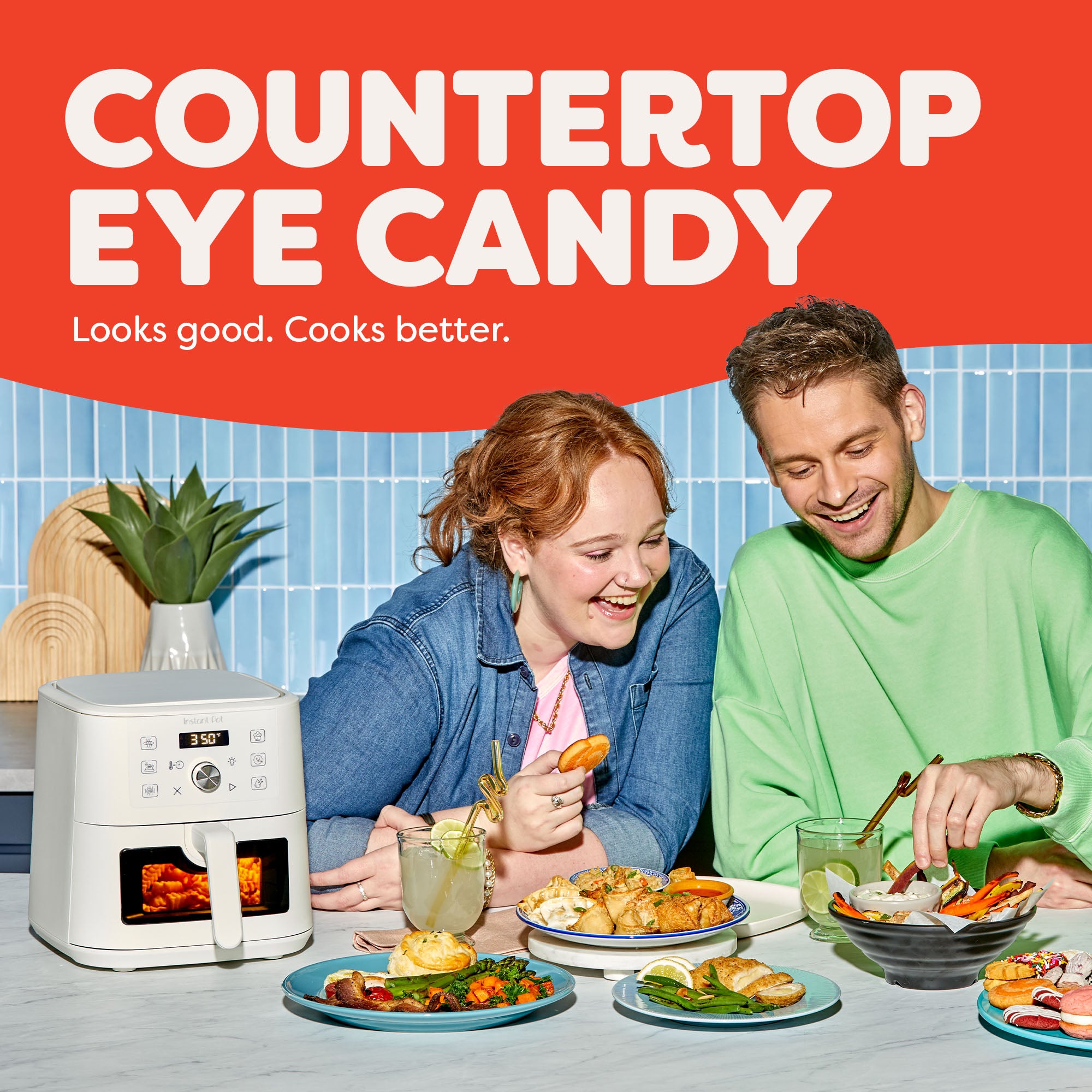 Two smiling people sit at a kitchen counter enjoying a variety of colorful meals, with the Instant Pot® 4QT Vortex® Plus Mini Air Fryer in Sea Salt on the counter beside them. The appliance shows a 350°F reading. Text above reads “Countertop Eye Candy – Looks good. Cooks better.” A potted plant and bright tiled backsplash complete the vibrant kitchen scene.
