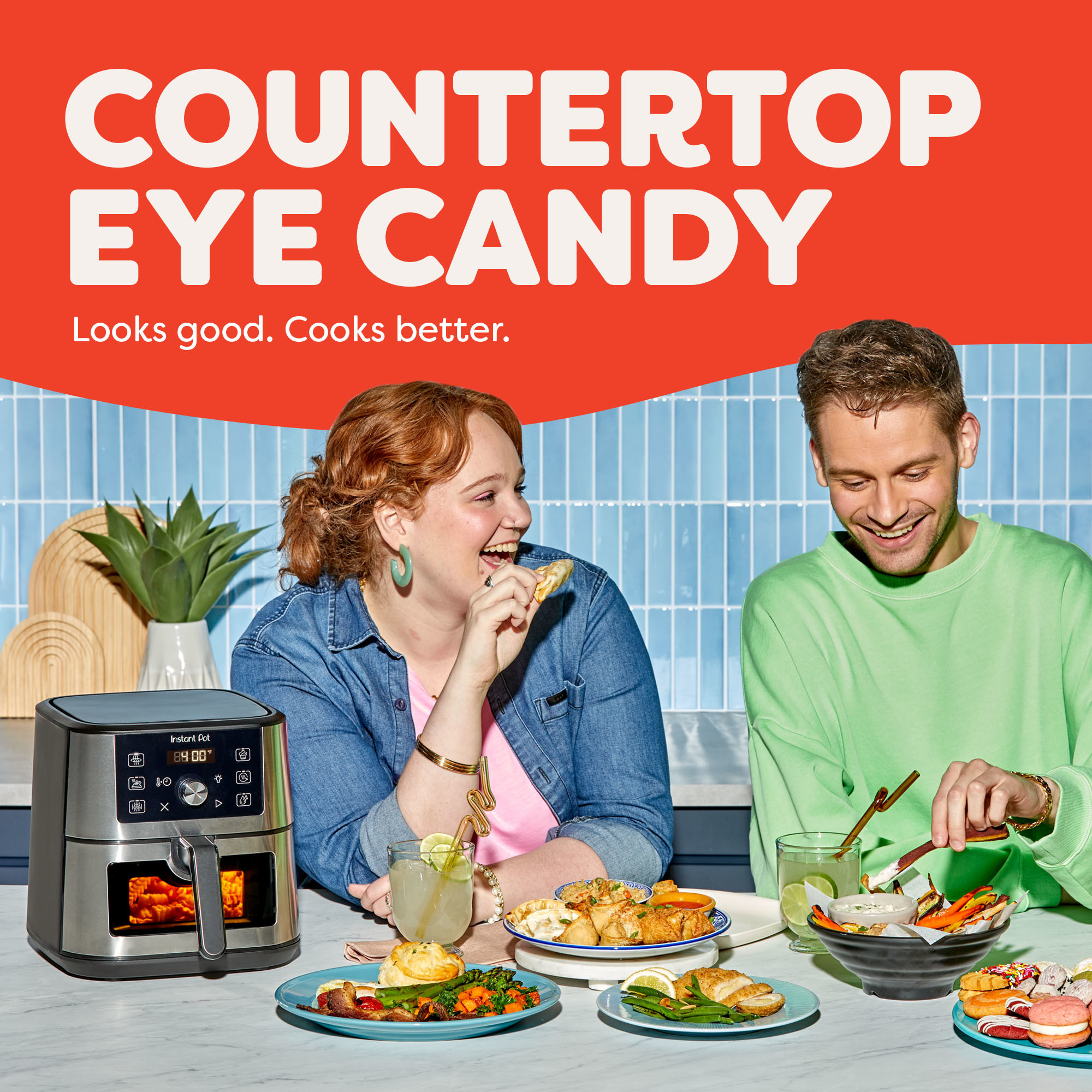 Two smiling people sit at a kitchen counter enjoying a variety of colorful meals, with the Instant Pot® 4QT VORTEX® Mini Air Fryer in Stainless Steel on the counter beside them. The appliance shows a 400°F reading. Text above reads “Countertop Eye Candy – Looks good. Cooks better.” A potted plant and bright tiled backsplash complete the vibrant kitchen scene.