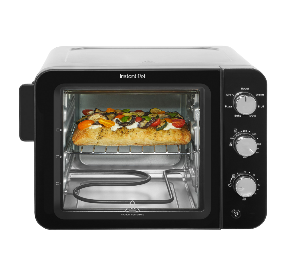 Front-facing view of the Instant Pot® 4-Slice InstantHeat™ Mini Air Fryer Toaster Oven with the door closed. A slice of focaccia bread topped with vegetables and cheese is baking on the middle rack. Manual control dials for temperature, timer, and cooking functions are on the right side of the unit.