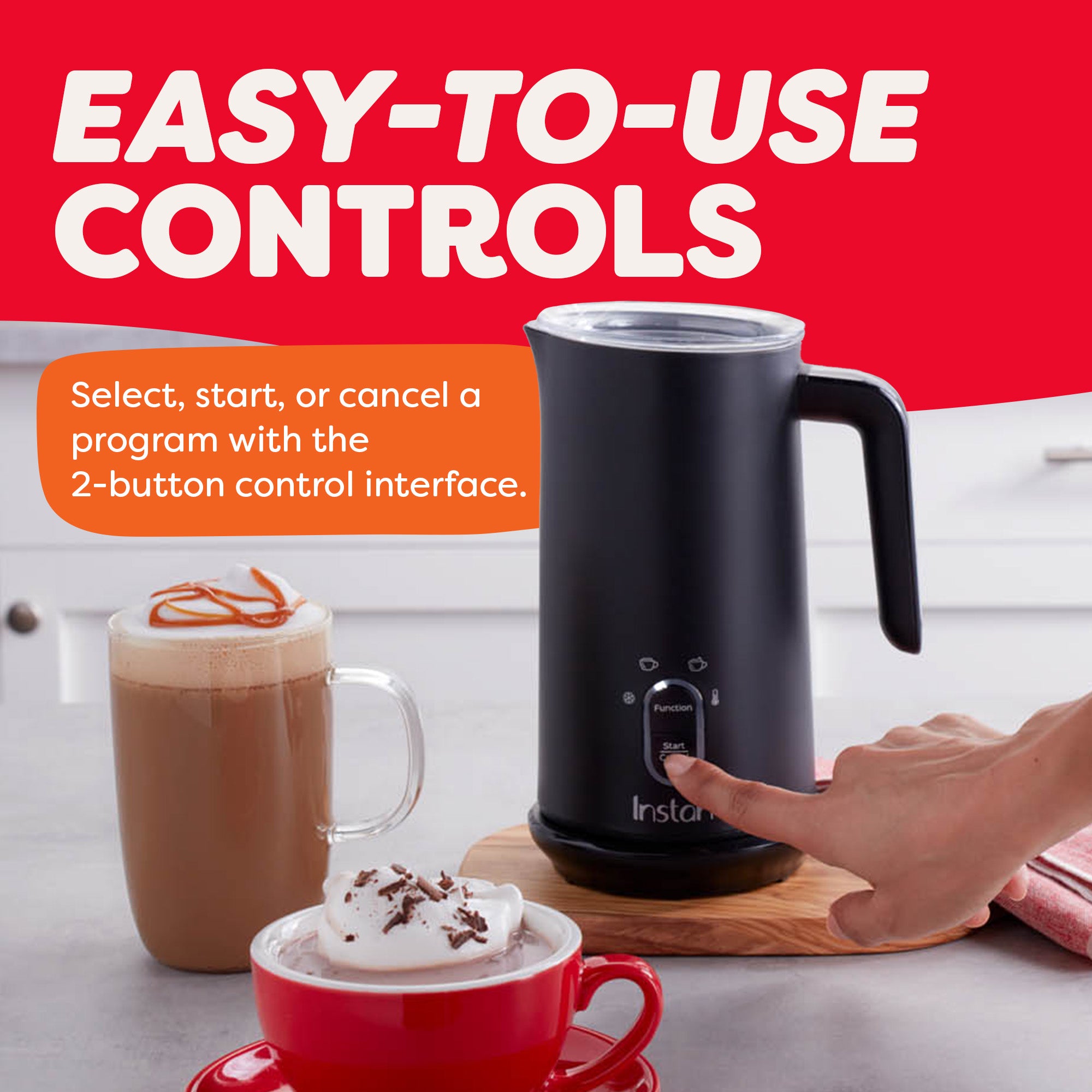 Finger pressing a button on a black Instant milk frother with two-button control interface. Text reads: 'Easy-to-use controls – Select, start, or cancel a program with the 2-button control interface.' A caramel latte in a glass mug and hot chocolate in a red ceramic mug are placed nearby on the kitchen counter.