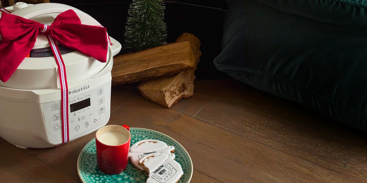 A Sea Salt–colored Instant Pot® 4-quart RIO™ Mini multi-cooker wrapped with a red ribbon sits on a wooden floor beside iced cookies and a glass of milk in a cozy holiday setting.