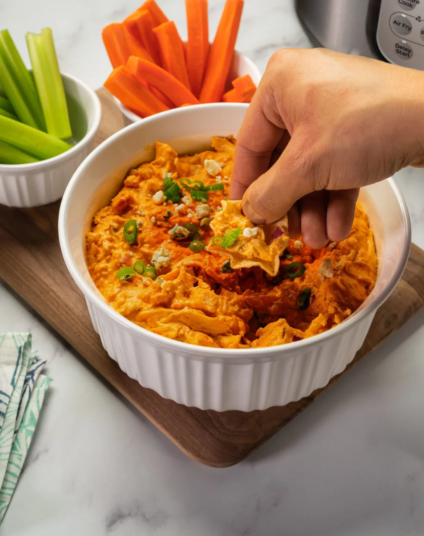 A hand dipping a tortilla chip into a creamy orange buffalo-style dip topped with sliced green onions and crumbled cheese, served in a white ramekin on a wooden board with celery and carrot sticks nearby.