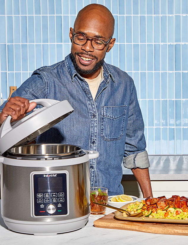 Man picking up a lid off an Instant Pot Multi-Cooker with Whisper Quiet functionality. In the foreground is a plate of sweet potatoes on top of a bed of corn with zucchini. 