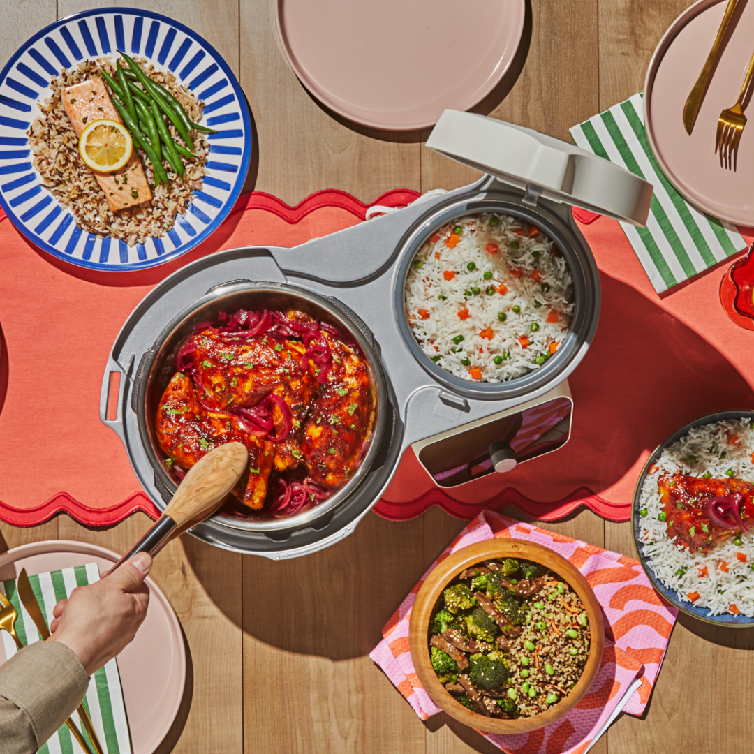 Overhead view of the Instant Pot® Dual Action Multi-Cooker in Sea Salt on a wooden table, with two open pots cooking simultaneously. The larger pot holds saucy chicken topped with pickled red onions as a hand stirs with a wooden spoon. The smaller pot contains white rice with peas and diced carrots. Surrounding the appliance are plated meals including salmon with green beans over rice, a bowl of broccoli and beef with grains, and additional rice dishes, styled with colorful plates and striped napkins