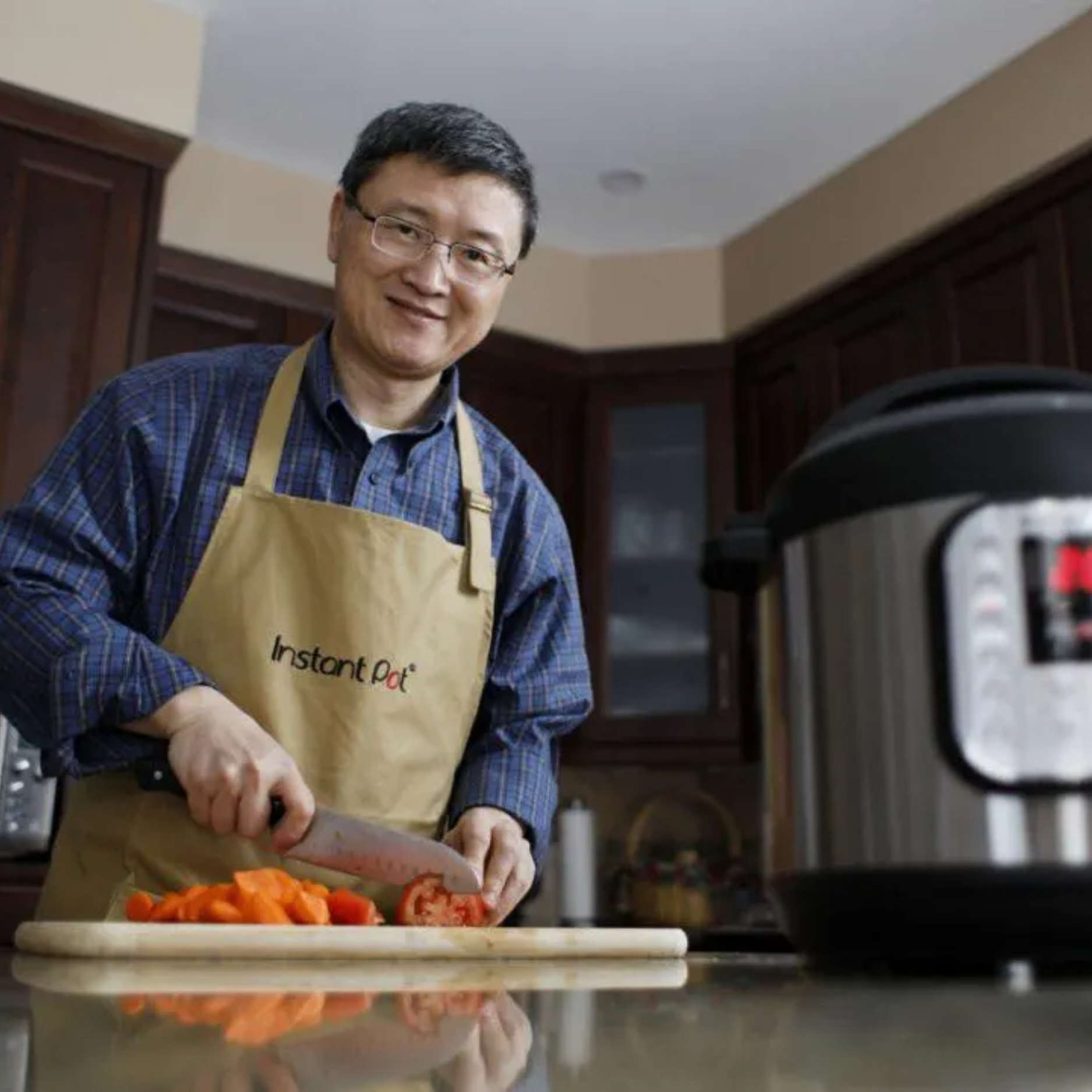  Dr. Robert Wang, co-founder of Instant Pot®, smiling as he slices tomatoes and bell peppers on a cutting board in a home kitchen. He’s wearing an Instant Pot® apron, with an Instant Pot® appliance visible in the foreground.