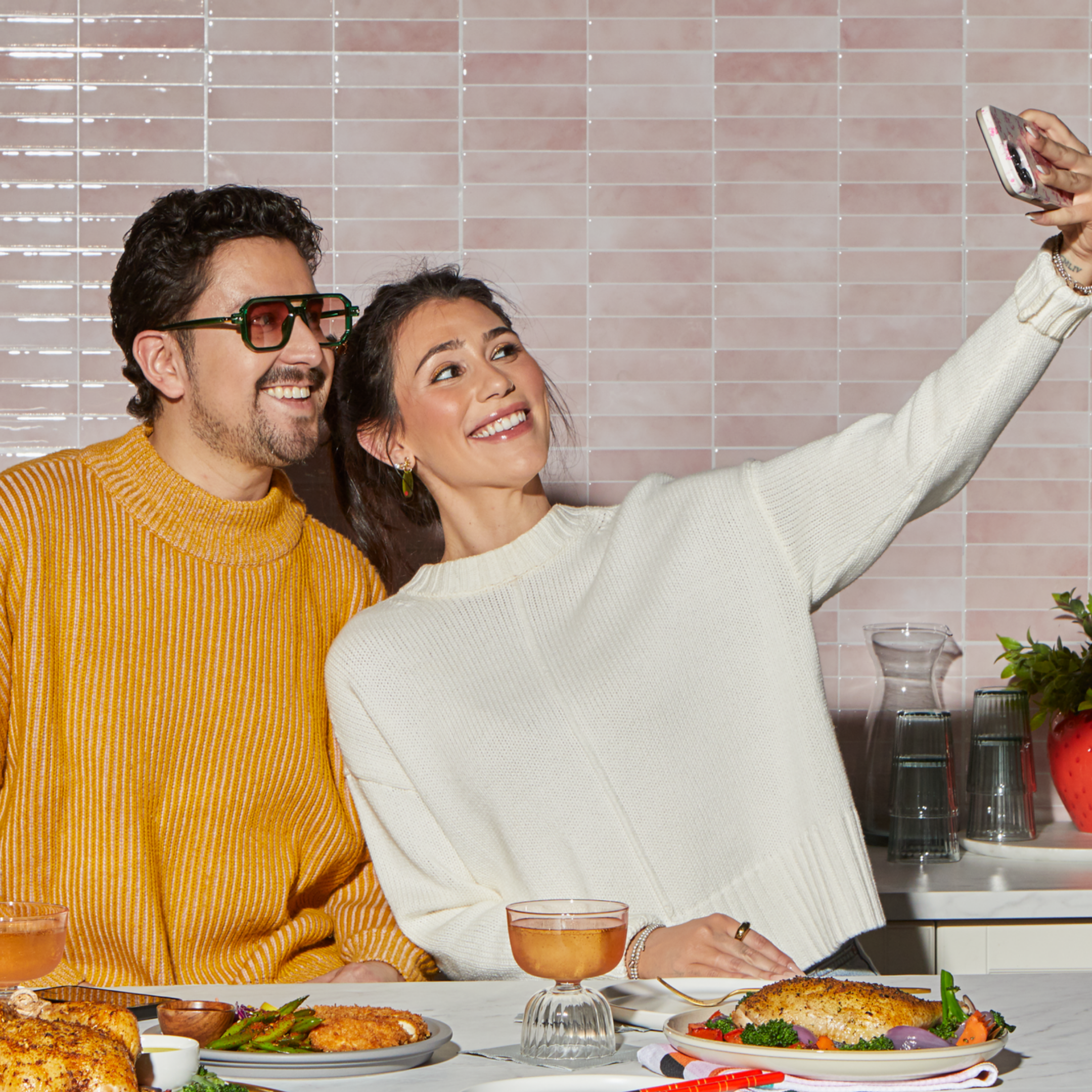 A couple smiles while taking a selfie at a kitchen counter, seated in front of plates filled with colorful meals and glasses of a light pink drink. One wears a mustard yellow sweater and large sunglasses, while the other wears an off-white sweater. Behind them is a soft pink tiled wall with stacked glassware and a vase of greenery.