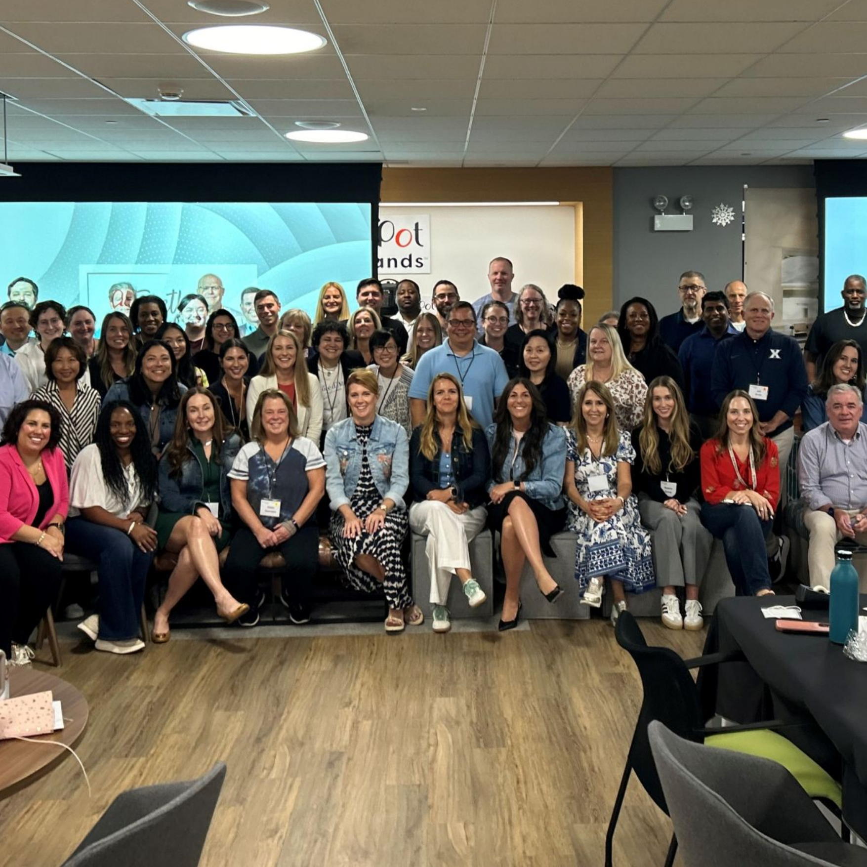 Large group of people smiling and posing for a group photo inside a conference or meeting room, with a presentation screen and Instant Brands signage in the background.