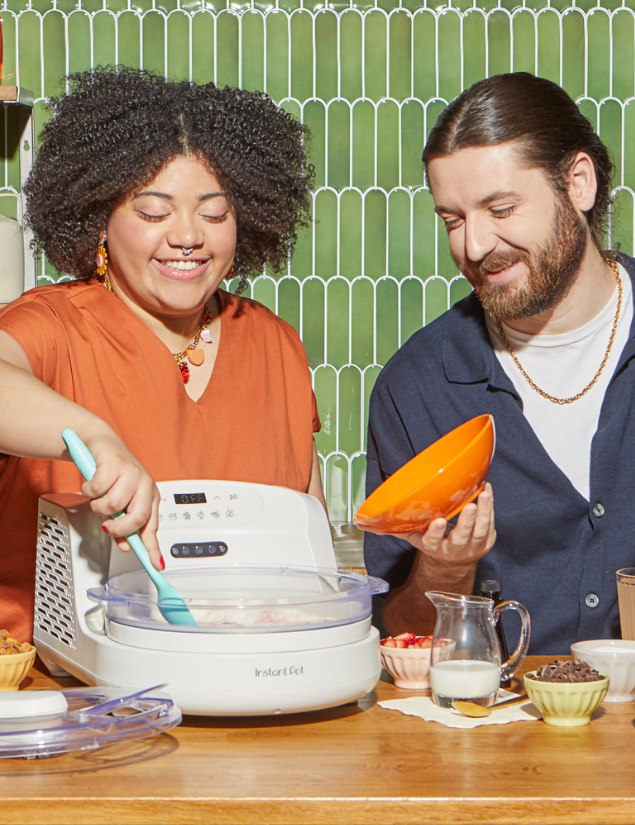 Two people smile while making ice cream with the Sea Salt InstantChill™ Ice Cream Maker on a wooden countertop, one stirring the mixture inside the open machine while the other holds an orange bowl. Surrounding them are ingredients like sliced strawberries, chocolate chips, and a small pitcher of milk, set against a green tiled backsplash.