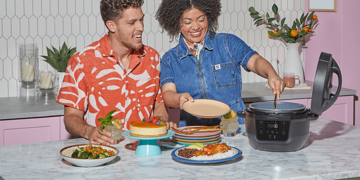 Two people smiling in a brightly lit kitchen as one serves food from a black Instant Pot® RIO™ Mini Multi-Cooker on the counter, surrounded by colorful plates, drinks, and vibrant dishes.