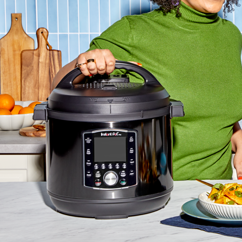 A person in a green sweater places their hand on the lid of a black Instant Pot Pro on a kitchen counter. The pot’s control panel is visible, showing multiple one-touch cooking programs. In the foreground, there is a bowl of food and a folded napkin, while in the background, a bowl of oranges and wooden cutting boards sit on the countertop.