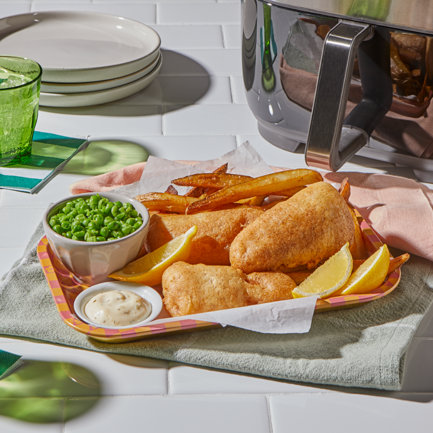 A tray of crispy fish and chips is served with lemon wedges, tartar sauce, and a side of green peas, placed on a checkered tray over folded kitchen towels. In the background, an Instant Pot Air Fryer is partially visible on a white tiled countertop, along with a green glass, striped napkin, and stacked plates
