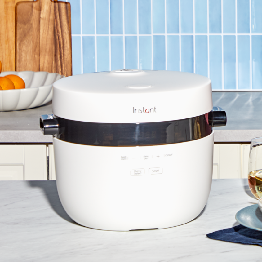 A white Instant Pot rice cooker sits on a kitchen counter in a brightly lit setting. The sleek, modern design features a minimalist control panel with buttons for cooking functions. In the background, a bowl of oranges and wooden cutting boards rest on the counter, while a glass of white wine and a plate are partially visible in the foreground.