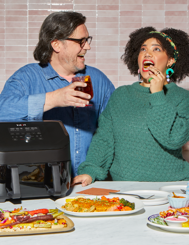 A man and woman share a laugh in a bright kitchen, standing behind a counter filled with colorful dishes including roasted vegetables and salmon. The woman, dressed in a green sweater, takes a bite of food while the man holds a drink. An Instant Pot® air fryer sits on the counter in front of them, with a pink tiled backsplash in the background.