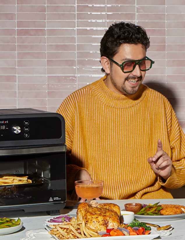 A man in a mustard yellow striped sweater and green sunglasses smiles while holding a drink and sitting at a table filled with a roasted chicken, fries, vegetables, and dipping sauces. An Instant Pot® toaster oven is beside him, mid-cook with fries inside, set against a glossy pink tiled wall.